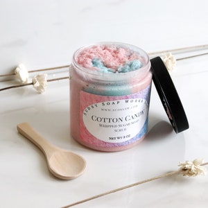 May include: A clear jar of Cotton Candy Whipped Sugar Soap Scrub with a black lid and a small wooden spoon. The scrub is pink and blue, and the label reads "Cotton Candy" and "Net Wt 8 oz."
