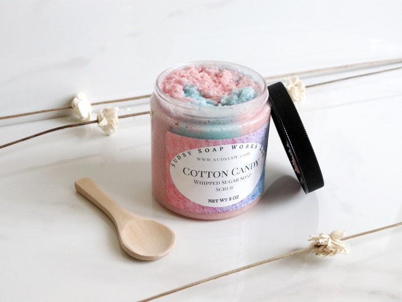 May include: A clear jar of Cotton Candy Whipped Sugar Soap Scrub with a black lid and a small wooden spoon. The scrub is pink and blue, and the label reads "Cotton Candy" and "Net Wt 8 oz."