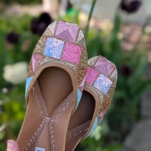 May include: A pair of embellished, flat shoes with a geometric pattern in pink, blue, and gold sequins. The shoes have a tan leather sole and the text "CHICAGO KHUSSA" on the insole. The background is a blurred garden scene.
