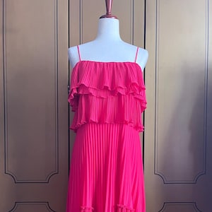 May include: A vibrant hot pink midi dress featuring a pleated texture throughout. The garment has thin spaghetti straps, a layered ruffled bodice, and a tiered skirt with a handkerchief hemline that falls below the knees on a mannequin.