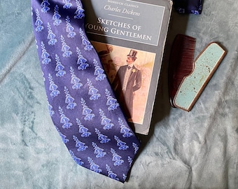 May include: A blue silk tie with a white floral pattern, a book titled "Sketches of Young Gentlemen" by Charles Dickens, a comb, and a small metal case with a razor.