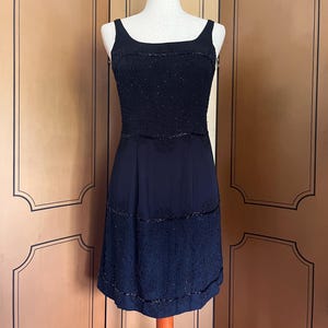 May include: A black sleeveless dress with a scoop neckline and a knee-length hemline. The bodice is heavily embellished with beads, and the skirt features beaded detailing. The dress is displayed on a mannequin against a neutral background.