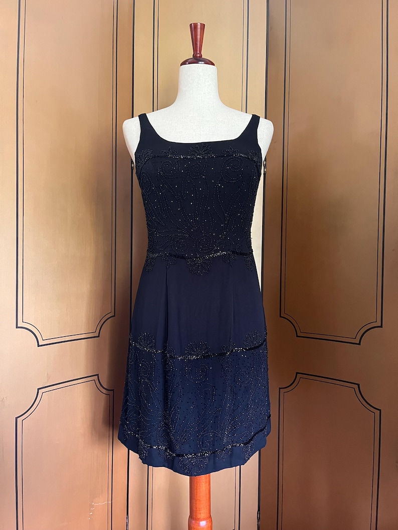 May include: A black sleeveless dress with a scoop neckline and a knee-length hemline. The bodice is heavily embellished with beads, and the skirt features beaded detailing. The dress is displayed on a mannequin against a neutral background.