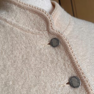 Op de afbeelding: Close-up van een lichtperzik jasje met een getextureerde gebreide stof. Het jasje heeft een ronde halslijn en is afgewerkt met een lichtere perzikgekleurde gebreide rand. Twee zilverkleurige knopen met een gedetailleerd ontwerp zijn zichtbaar.