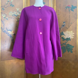 May include: A vibrant purple coat with three bright pink buttons and a single pocket. The coat is displayed on a mannequin, showcasing its tailored design and long sleeves. The coat's color is a rich, saturated hue.
