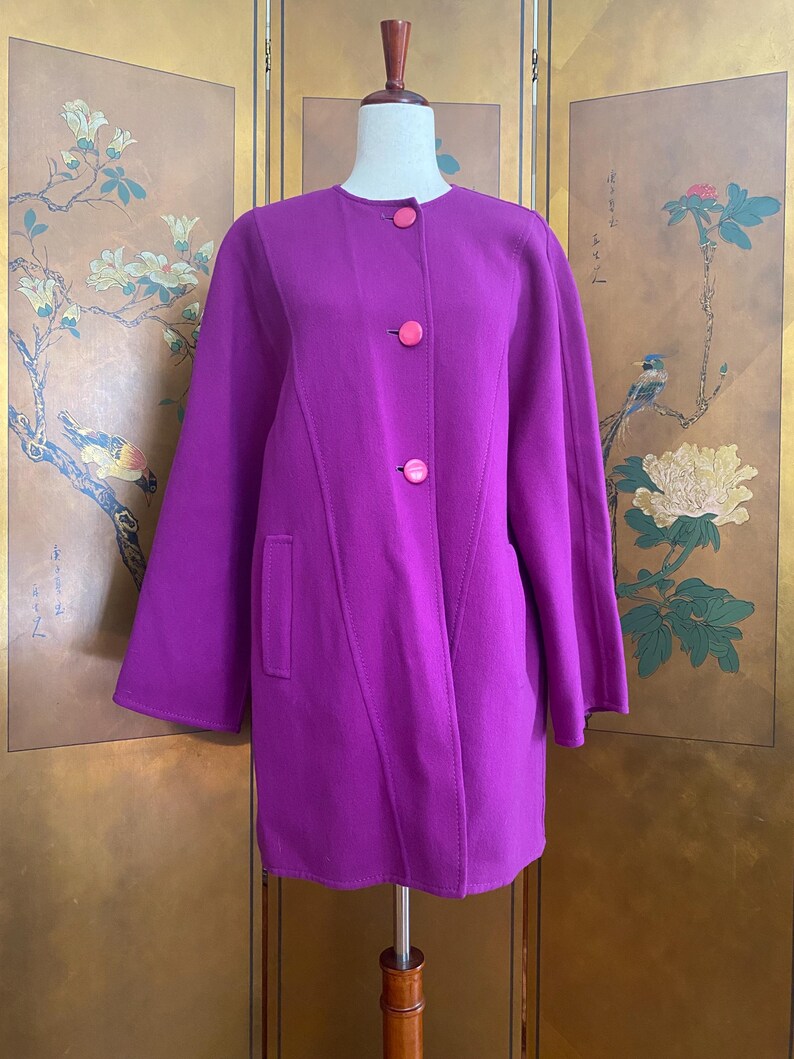 May include: A vibrant purple coat with three bright pink buttons and a single pocket. The coat is displayed on a mannequin, showcasing its tailored design and long sleeves. The coat's color is a rich, saturated hue.
