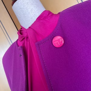 May include: A close-up of a two-tone garment on a mannequin. The outer layer is a deep purple, with a large, round, pink button. Underneath is a bright pink blouse. The garment is likely a jacket or blazer.