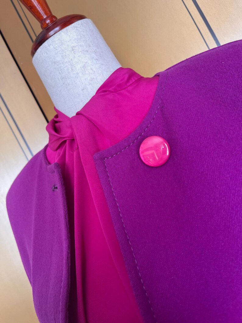 May include: A close-up of a two-tone garment on a mannequin. The outer layer is a deep purple, with a large, round, pink button. Underneath is a bright pink blouse. The garment is likely a jacket or blazer.