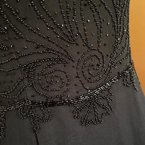 May include: Close-up of a black dress bodice with intricate beadwork. The design features swirling patterns and linear accents, creating a textured and elegant look. The dress appears to be made of a soft, flowing fabric.