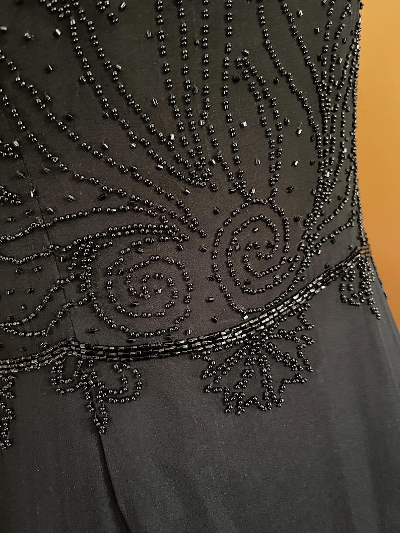 May include: Close-up of a black dress bodice with intricate beadwork. The design features swirling patterns and linear accents, creating a textured and elegant look. The dress appears to be made of a soft, flowing fabric.