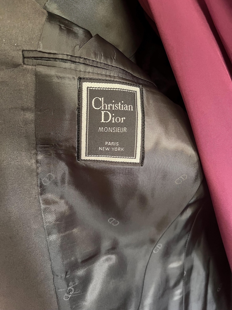 Pu&ograve; includere: Primo piano dell'interno di un blazer Christian Dior Monsieur grigio scuro. L'etichetta recita "Christian Dior MONSIEUR PARIS NEW YORK" in una cornice bianca e nera. La fodera &egrave; grigio scuro con un sottile motivo "CD".