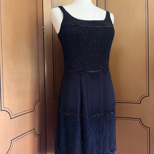 May include: A black sleeveless dress with intricate beaded embellishments. The dress features a square neckline and a fitted bodice with detailed patterns. The skirt has a subtle A-line shape, and the beading adds texture and visual interest to the garment.