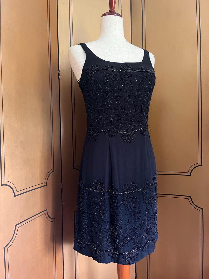 May include: A black sleeveless dress with intricate beaded embellishments. The dress features a square neckline and a fitted bodice with detailed patterns. The skirt has a subtle A-line shape, and the beading adds texture and visual interest to the garment.