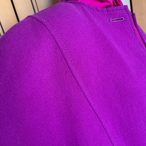 May include: Close-up of a vibrant magenta jacket with a tailored design. The jacket features a single button closure and a contrasting fuchsia lining visible at the neckline. The stitching details and fabric texture are clearly visible.