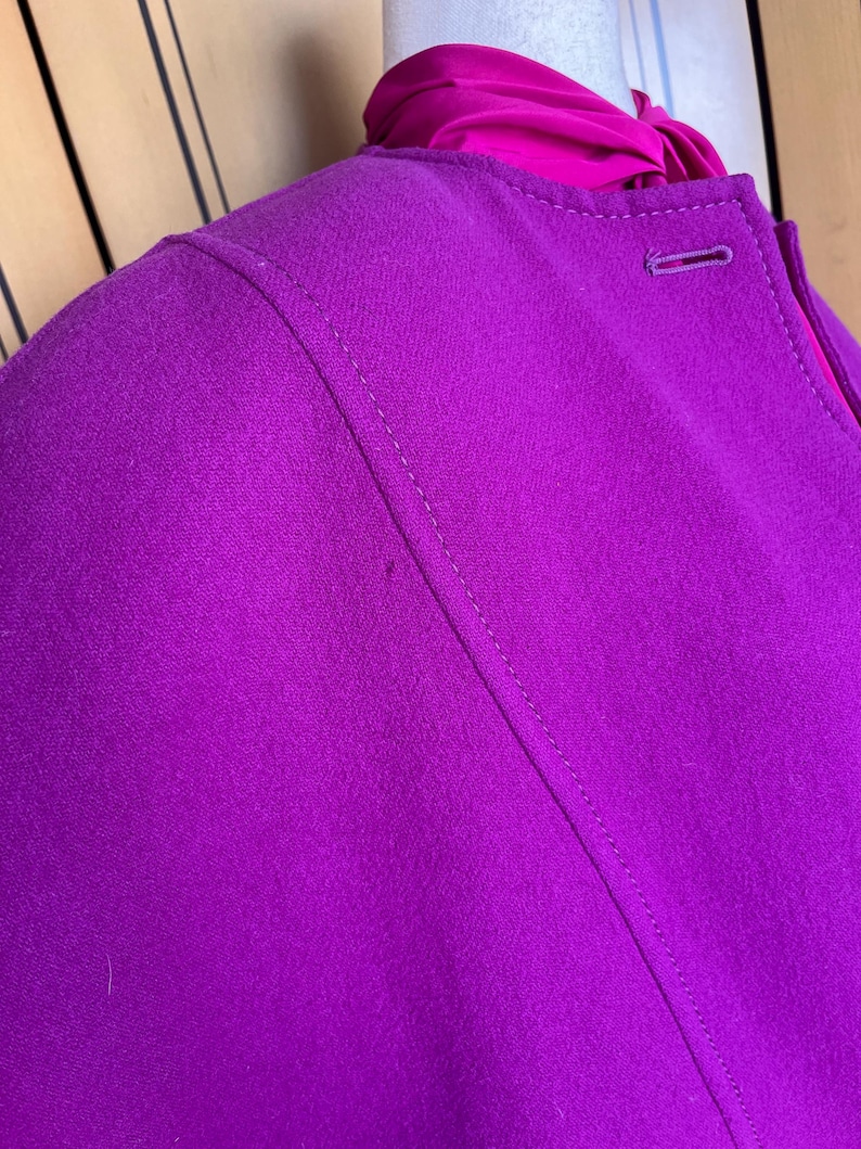 May include: Close-up of a vibrant magenta jacket with a tailored design. The jacket features a single button closure and a contrasting fuchsia lining visible at the neckline. The stitching details and fabric texture are clearly visible.