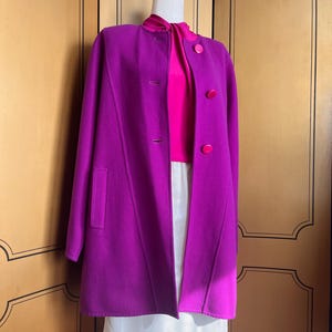 May include: A vibrant magenta coat with a matching magenta top and large buttons. The coat has a classic design with a high collar and a tailored fit. The outfit is displayed on a mannequin, showcasing the coat's elegant silhouette and color.