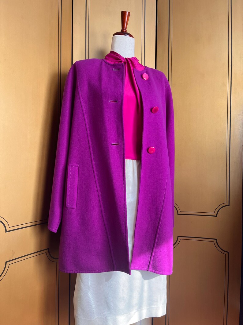 May include: A vibrant magenta coat with a matching magenta top and large buttons. The coat has a classic design with a high collar and a tailored fit. The outfit is displayed on a mannequin, showcasing the coat's elegant silhouette and color.