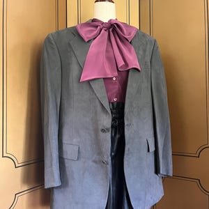 Pu&ograve; includere: Un blazer grigio con un papillon bordeaux e una camicia abbinata. Il blazer ha due bottoni e due tasche. L'outfit &egrave; esposto su un manichino.