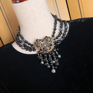 May include: A vintage black beaded necklace with a gold-toned centerpiece adorned with iridescent rhinestones. The necklace features multiple strands of black beads and a cascading fringe of smaller beads, displayed on a mannequin.