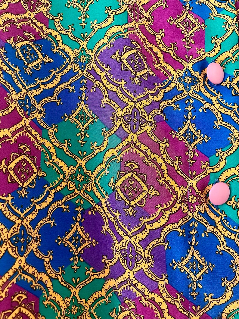 May include: A close-up of a fabric with a colorful geometric pattern. The fabric is a mix of blue, purple, green, and yellow. The pattern is made up of repeating squares and diamonds. There are two pink buttons on the fabric.