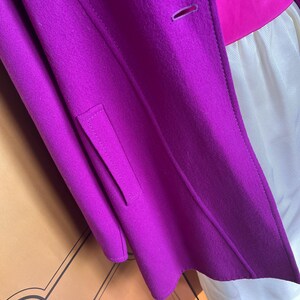 May include: A close-up of a vibrant purple coat with a tailored design. The coat features a single button closure and a small pocket detail. The coat is layered over a pink top and a white garment.