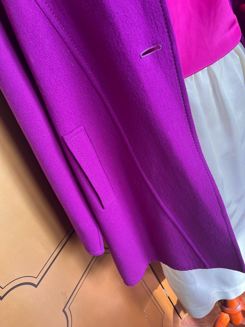 May include: A close-up of a vibrant purple coat with a tailored design. The coat features a single button closure and a small pocket detail. The coat is layered over a pink top and a white garment.