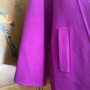 May include: A close-up of a vibrant purple coat, showcasing the sleeve and a pocket detail. The coat's fabric has a soft texture, and the stitching is visible. The design is simple and elegant, with a focus on the rich color.
