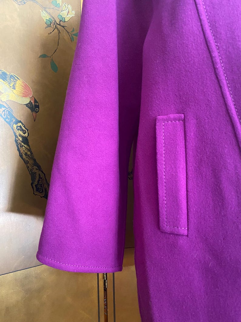 May include: A close-up of a vibrant purple coat, showcasing the sleeve and a pocket detail. The coat's fabric has a soft texture, and the stitching is visible. The design is simple and elegant, with a focus on the rich color.
