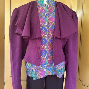 May include: A purple blazer with a ruffled collar and long sleeves. The blazer is worn over a patterned blouse with a ruffled bottom. The blouse has a geometric pattern in shades of pink, blue, and gold.