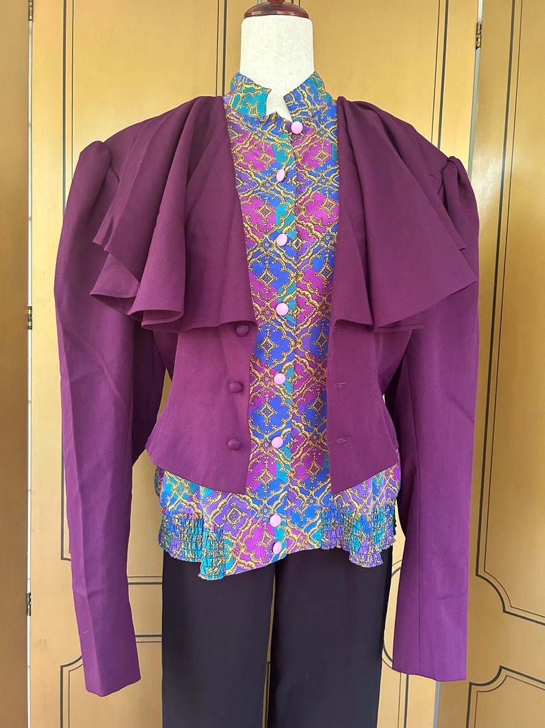 May include: A purple blazer with a ruffled collar and long sleeves. The blazer is worn over a patterned blouse with a ruffled bottom. The blouse has a geometric pattern in shades of pink, blue, and gold.