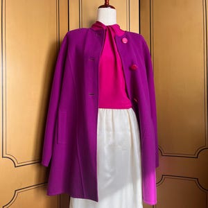 May include: A vibrant purple coat with a matching pink blouse and a white skirt. The coat features a classic design with buttons and pockets. The outfit is displayed on a mannequin against a wooden background.