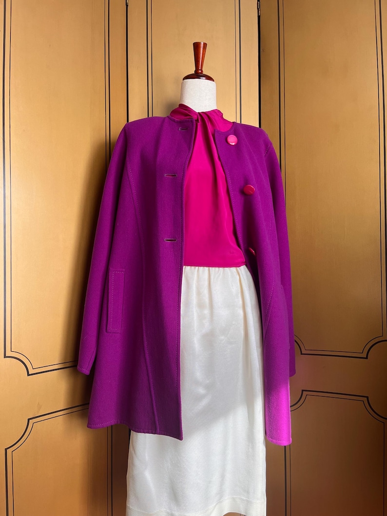 May include: A vibrant purple coat with a matching pink blouse and a white skirt. The coat features a classic design with buttons and pockets. The outfit is displayed on a mannequin against a wooden background.