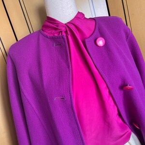 May include: A vibrant purple coat with a round collar and matching buttons. Beneath the coat, a bright pink blouse is visible. The outfit is displayed on a mannequin, showcasing a classic and colorful ensemble.