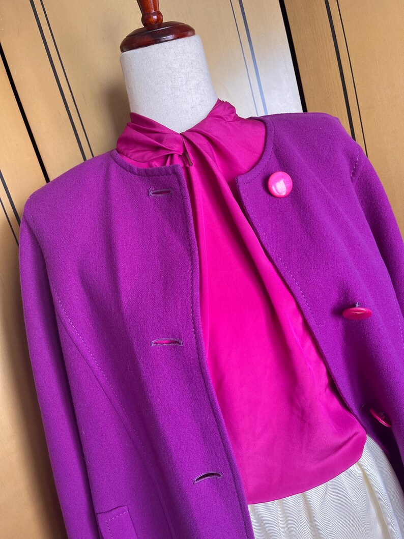 May include: A vibrant purple coat with a round collar and matching buttons. Beneath the coat, a bright pink blouse is visible. The outfit is displayed on a mannequin, showcasing a classic and colorful ensemble.