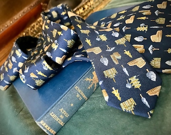 Puede incluir: Dos corbatas de seda azul oscuro con un patrón dorado y blanco de árboles y edificios estilizados. Las corbatas están sobre un libro azul con el título "Twilight in Italy" y el autor "D.H. Lawrence".