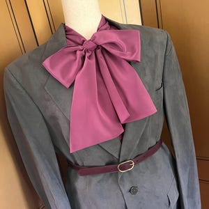 Pu&ograve; includere: Un blazer grigio con un grande papillon magenta, una cintura abbinata con fibbia dorata e una camicia con colletto magenta. Il blazer &egrave; abbottonato e ha tasche.