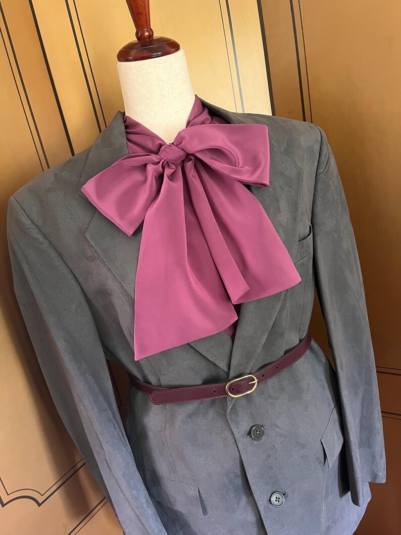 Pu&ograve; includere: Un blazer grigio con un grande papillon magenta, una cintura abbinata con fibbia dorata e una camicia con colletto magenta. Il blazer &egrave; abbottonato e ha tasche.