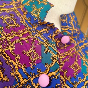 May include: A close-up of a blue, purple, and teal patterned fabric with gold accents. Two pink buttons are visible on the fabric.