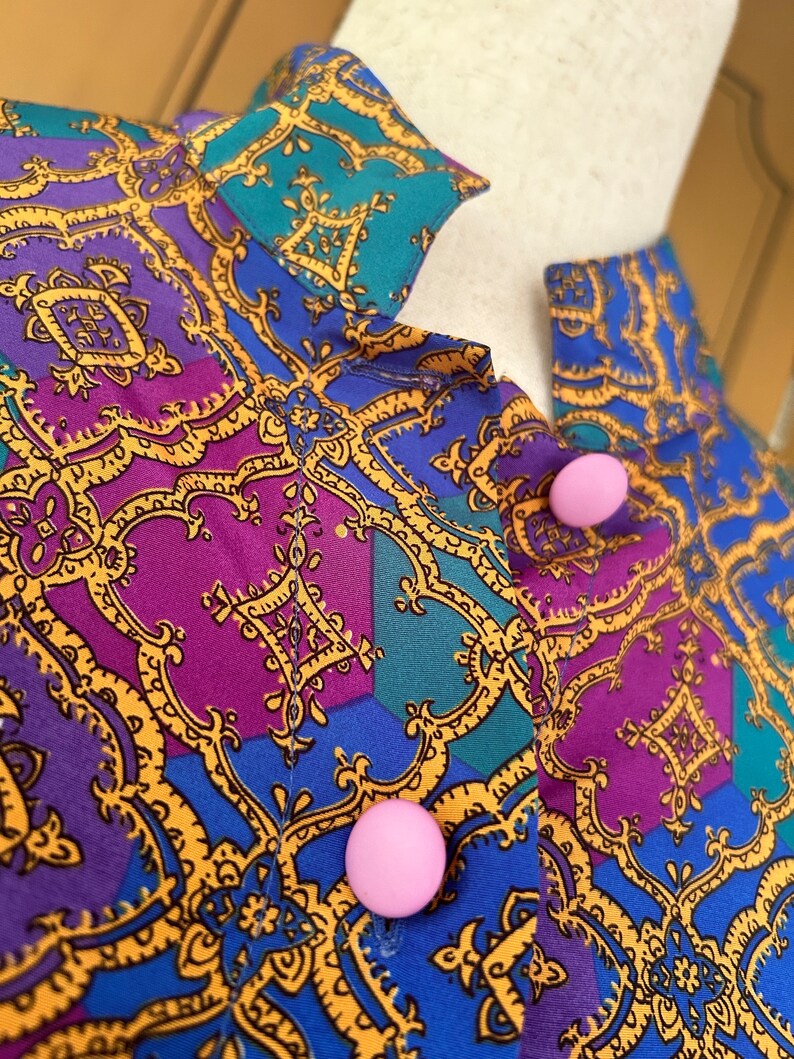 May include: A close-up of a blue, purple, and teal patterned fabric with gold accents. Two pink buttons are visible on the fabric.