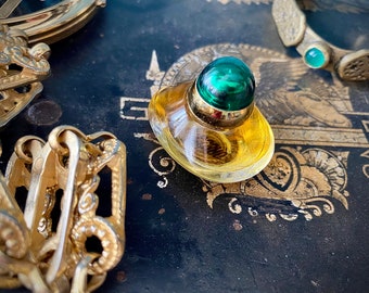 May include: A small, vintage perfume bottle with a green, spherical stopper and gold accents. The bottle contains a golden liquid. Other gold-toned decorative items are in the background, set against a dark, patterned surface.