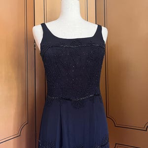 May include: A black evening gown with a square neckline and beaded embellishments. The bodice features intricate beadwork, and the skirt has a flowing design. The dress is displayed on a mannequin.