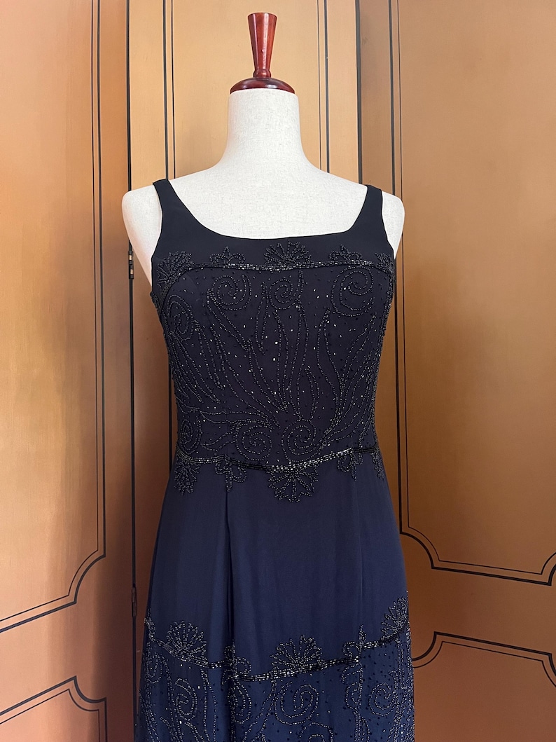 May include: A black evening gown with a square neckline and beaded embellishments. The bodice features intricate beadwork, and the skirt has a flowing design. The dress is displayed on a mannequin.