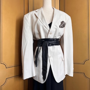 Puede incluir: Una blazer color crema con un cinturón de cuero negro que ciñe la cintura. Se ve una parte superior negra debajo de la blazer, y un pañuelo de bolsillo estampado añade detalles. La blazer se exhibe en un maniquí.