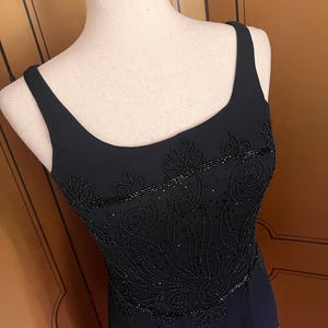 May include: A black evening dress with a scoop neckline and thin straps. The bodice is embellished with intricate beadwork in a floral design. The dress is displayed on a mannequin, showcasing its elegant silhouette and detailing.