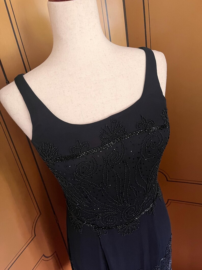 May include: A black evening dress with a scoop neckline and thin straps. The bodice is embellished with intricate beadwork in a floral design. The dress is displayed on a mannequin, showcasing its elegant silhouette and detailing.