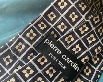 May include: Black and white silk tie with a geometric pattern of small white flowers in squares. The tie has a black label that reads "Pierre Cardin Pure Silk".