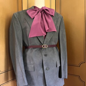 Pu&ograve; includere: Un blazer grigio con un papillon e una cintura bordeaux, esposto su un manichino. Il blazer ha una chiusura a due bottoni e tasche anteriori. La cintura ha una fibbia color oro.