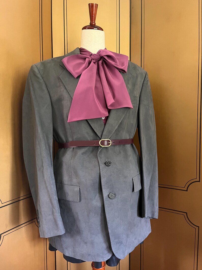 Pu&ograve; includere: Un blazer grigio con un papillon e una cintura bordeaux, esposto su un manichino. Il blazer ha una chiusura a due bottoni e tasche anteriori. La cintura ha una fibbia color oro.