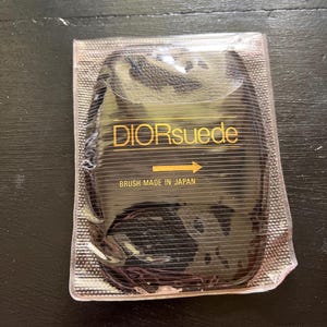 Pu&ograve; includere: Una spazzola Dior in camoscio nero in una confezione di plastica trasparente. Il testo "DIORsuede" &egrave; in lettere dorate, con una freccia che punta verso destra. Sotto c'&egrave; scritto "BRUSH MADE IN JAPAN".