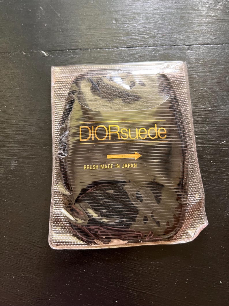 Pu&ograve; includere: Una spazzola Dior in camoscio nero in una confezione di plastica trasparente. Il testo "DIORsuede" &egrave; in lettere dorate, con una freccia che punta verso destra. Sotto c'&egrave; scritto "BRUSH MADE IN JAPAN".
